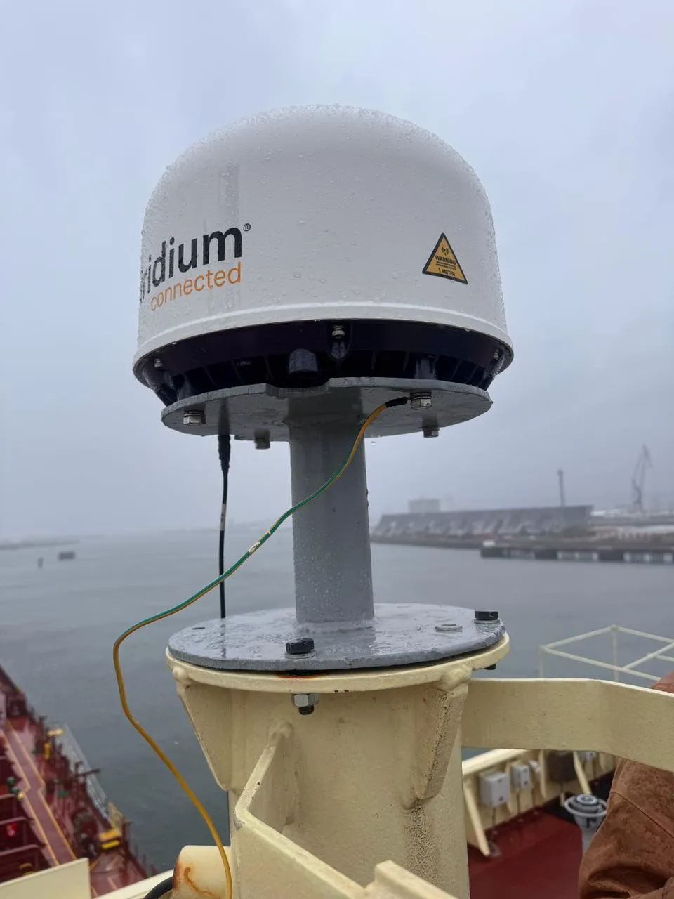 Iridium maritime antenna bolted to a mast pedestal on a tanker, earthing cable fitted, port in the background